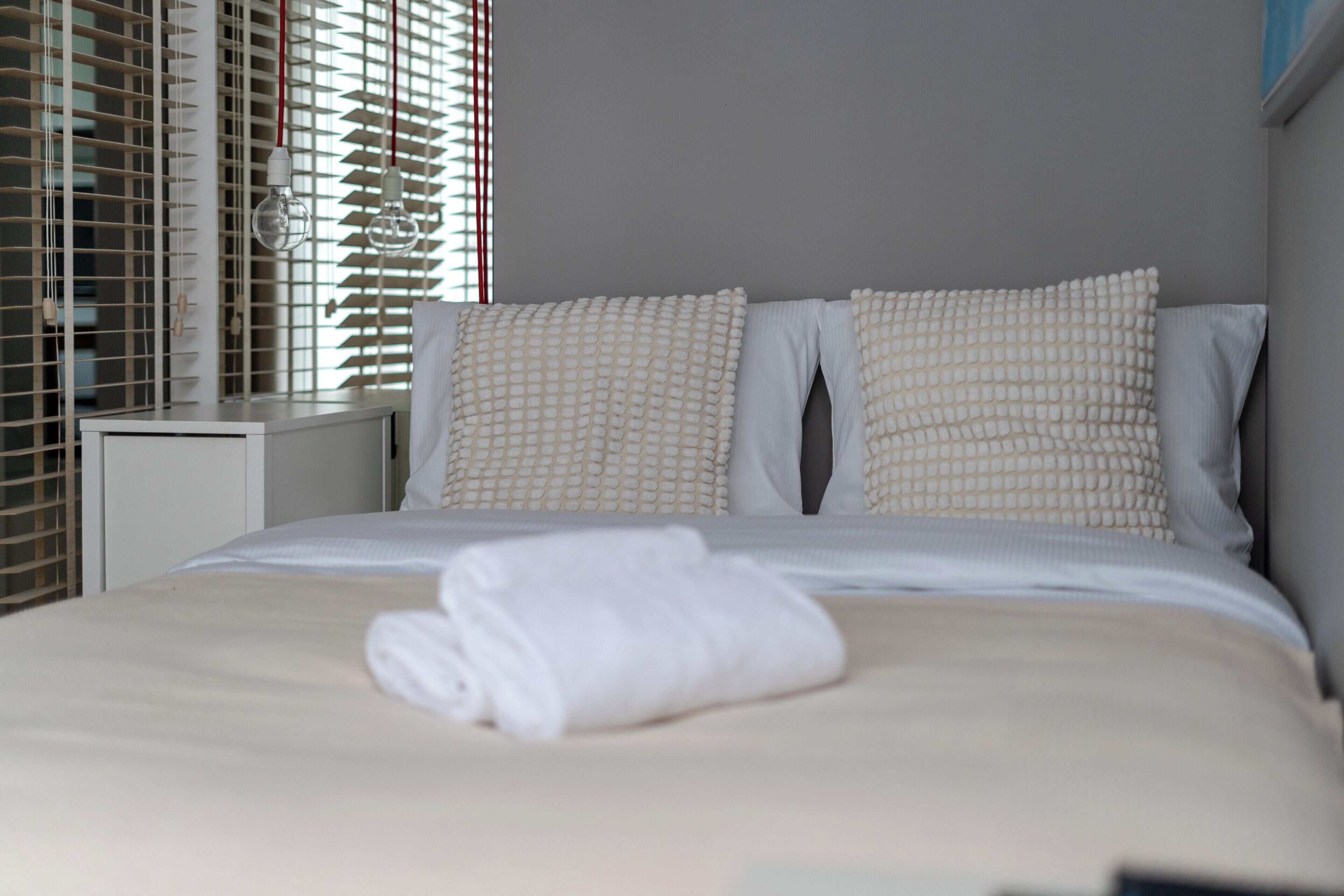 Uma cama bem arrumada com roupa de cama e almofadas brancas, uma toalha branca dobrada e uma mesa de cabeceira com abajur.