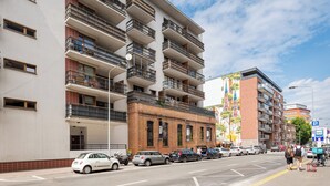 A multi-story building with balconies, a parking lot, and a street with cars and pedestrians.