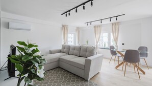 Ein modernes Wohnzimmer mit einem grauen Ecksofa, einem Glastisch mit vier Stühlen, einem gemusterten Teppich und einem großen Fenster mit Vorhängen.
