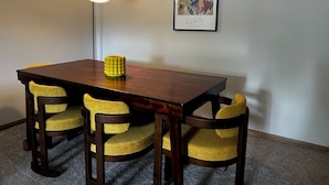 Un coin repas avec une table en bois, quatre chaises à l’assise rembourrée de couleur jaune, une lampe jaune et un tableau abstrait encadré accroché au mur.