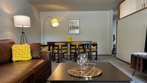 Un salon moderne avec un canapé en cuir marron, une table basse en bois sur laquelle sont posés des verres à vin, des fauteuils aux coussins jaunes et une table à manger.