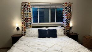 Un lit bien fait, avec une literie blanche et un oreiller bleu, flanqué de deux tables de chevet avec des lampes, une fenêtre avec des rideaux et un panier en osier.