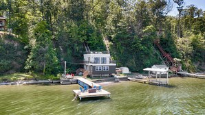 Une maison flottante dotée d’une terrasse et de sièges extérieurs, entourée d’une zone boisée et d’un quai.