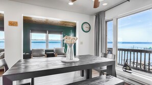 Un coin repas comprenant une table, des chaises, un vase de fleurs, une horloge et une vue sur l’eau grâce à de grandes fenêtres.