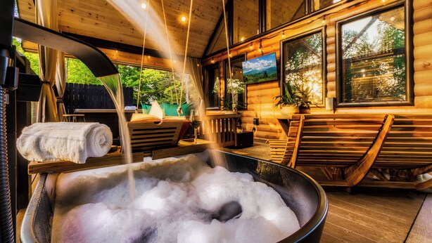 A hot tub with water flowing into it, wooden benches, and large windows with a view of greenery outside.