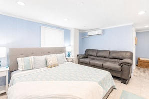 A bedroom with a bed, a grey sofa, an air conditioning unit, and a window with blinds.