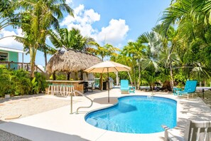 A swimming pool with lounge chairs and a bar area, surrounded by palm trees and a thatched-roof structure. - Private Florida Keys Escape w/ Pool, Dock, Tiki Bar – Pet Friendly (Little Torch Key)