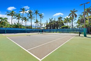 Een tennisbaan buiten met een net, omringd door palmbomen en een helderblauwe lucht.