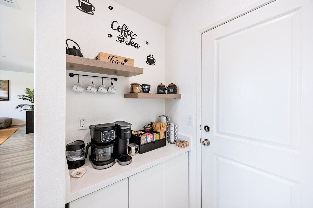Una kitchenette con cafetera, hervidor de agua y tazas para té en la pared, y estantes con frascos y recipientes.