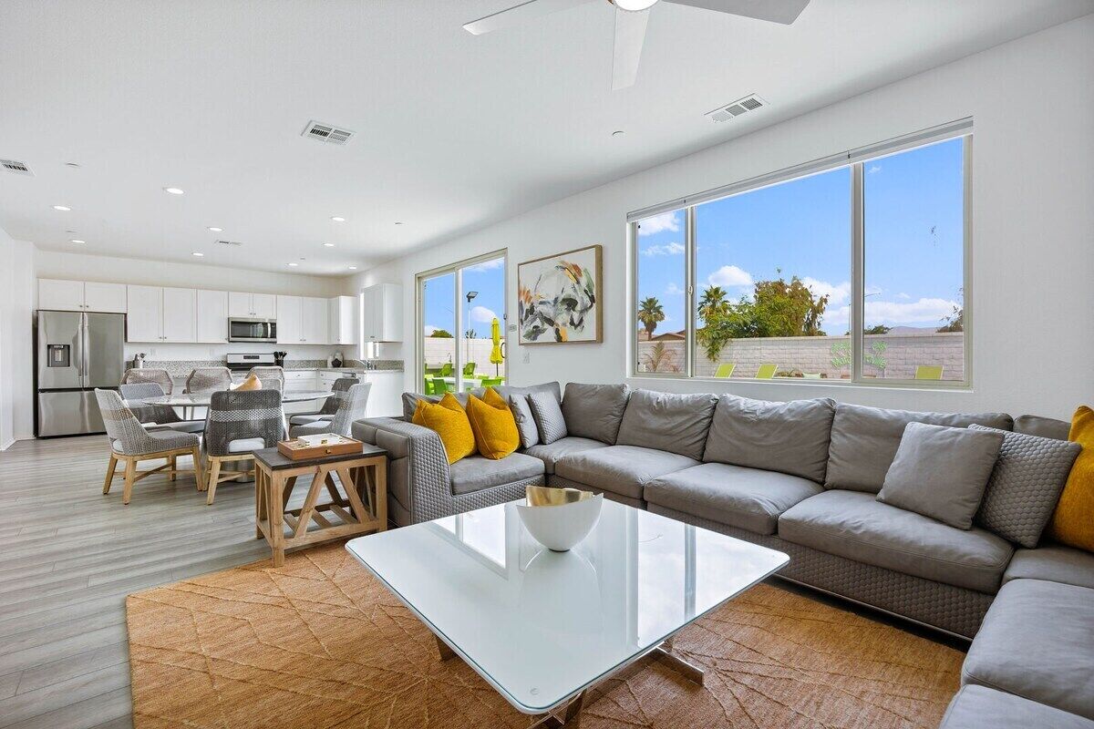 A modern living room with a glass coffee table, a grey sofa, and a dining area with chairs and a table.