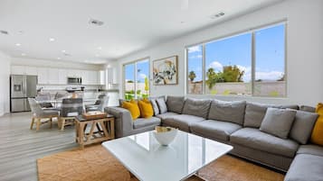 A modern living room with a glass coffee table, a grey sofa, and a dining area with chairs and a table.
