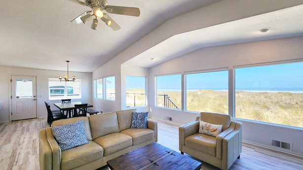 A spacious living room with a large window overlooking the beach, a ceiling fan, and a dining area with a chandelier. - Ocean Front on the Beach! Dog Friendly! (Waldport)
