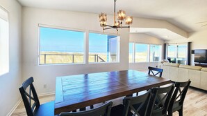 A spacious dining area with a large wooden table and chairs, large windows with a view of the beach, and a modern kitchen with a flat-screen TV. - Ocean Front on the Beach! Dog Friendly! (Waldport)