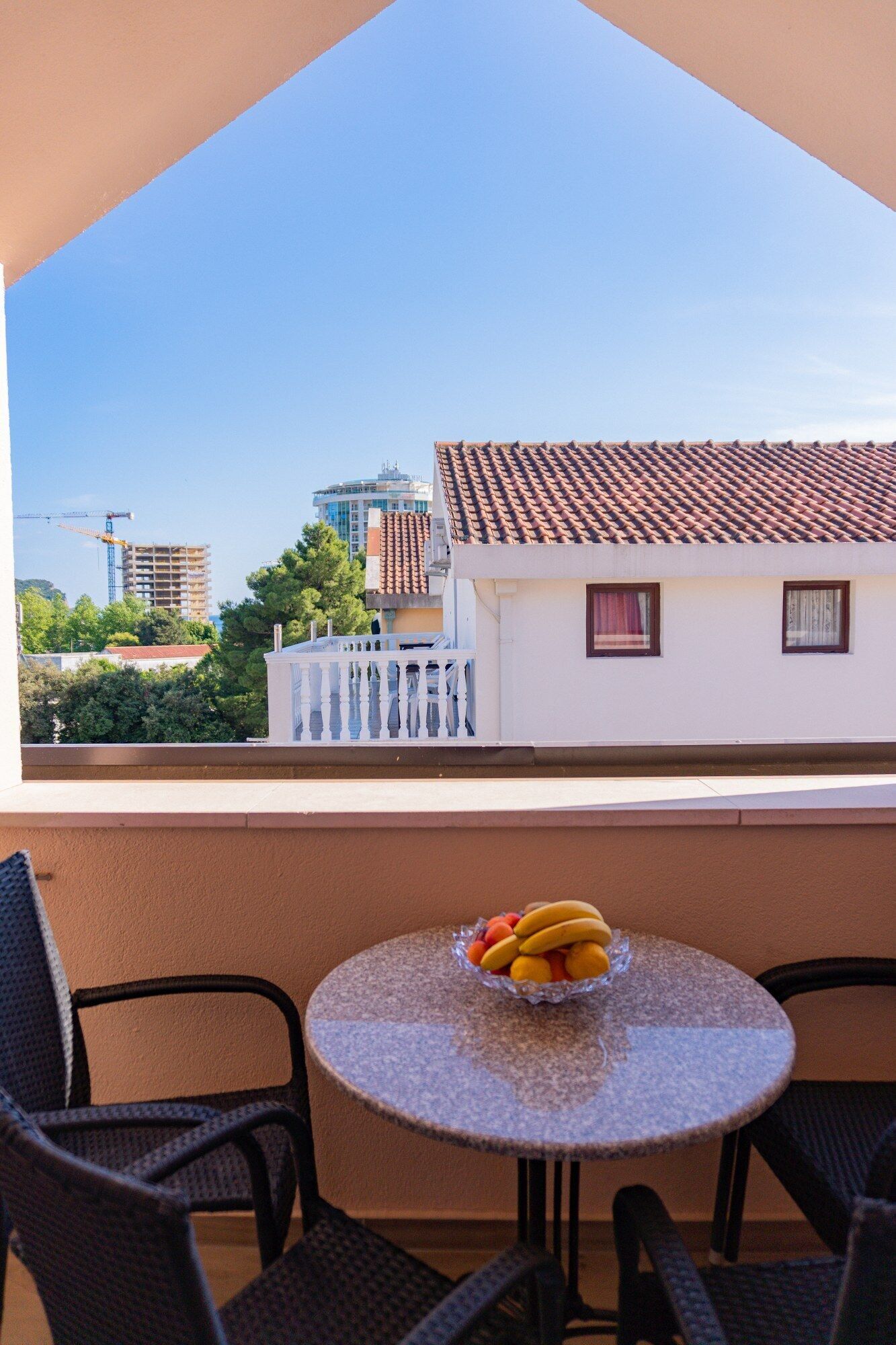 Un balcón con una mesa redonda y sillas, un cuenco de fruta y vistas a edificios y árboles.