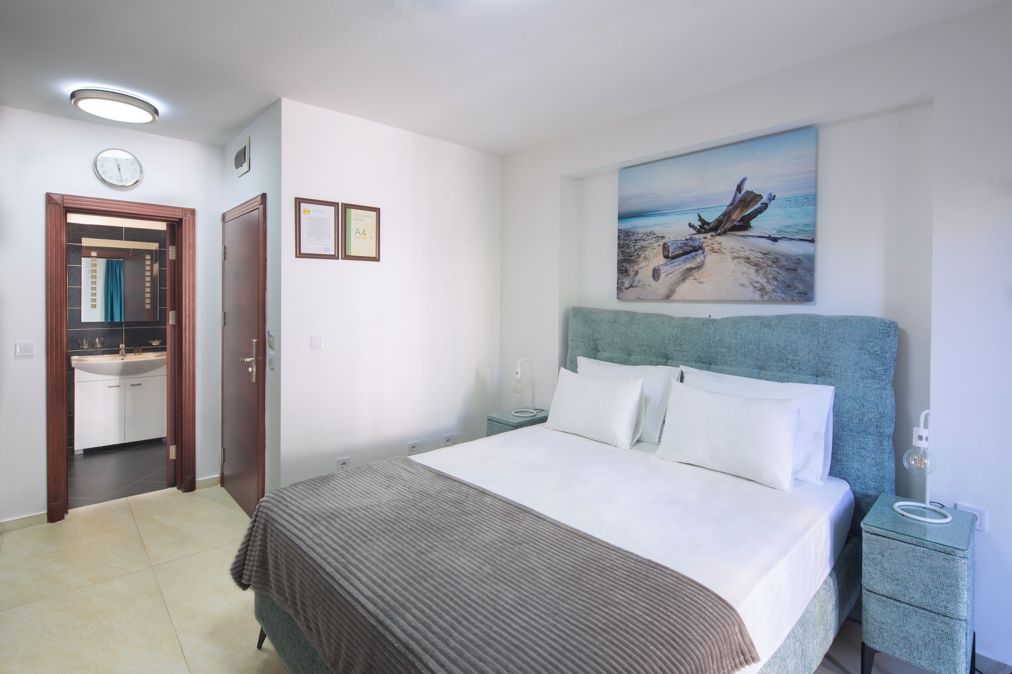 A bedroom with a bed, a nightstand, a painting of a beach scene, a clock, and a door leading to a bathroom.