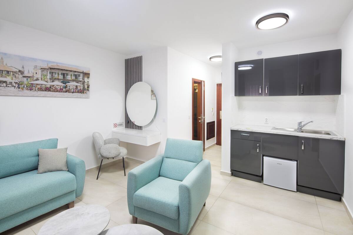 Un salon moderne avec un canapé, un fauteuil et un grand miroir rond. On y trouve également un coin cuisine doté de meubles sombres et d’un évier.