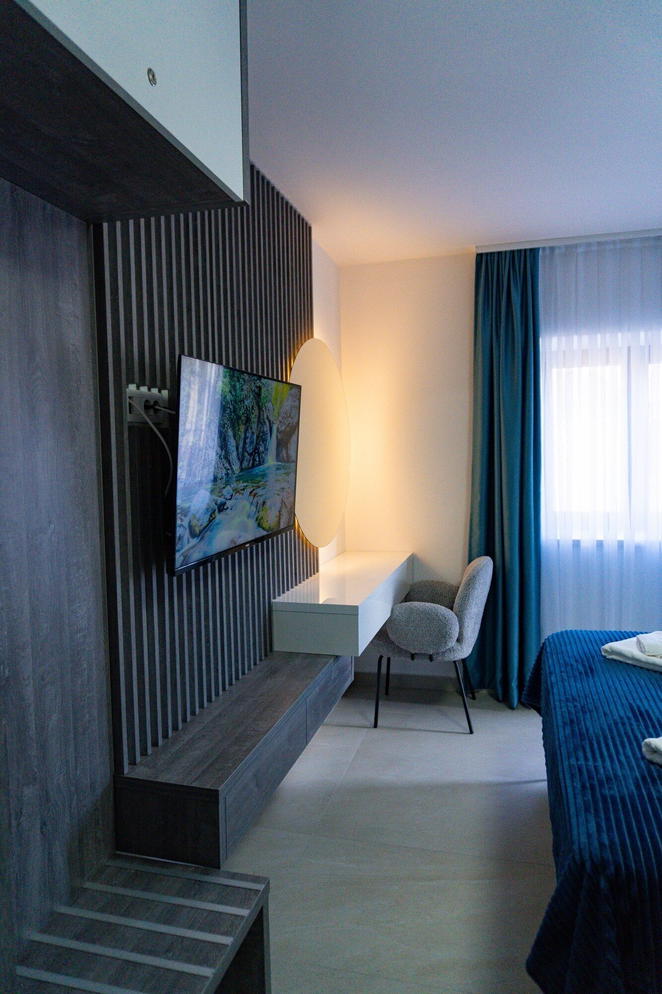 A modern hotel room with a flat-screen TV mounted on a wall, a desk with a chair, a bed with a blue bedspread, and a large window with curtains.