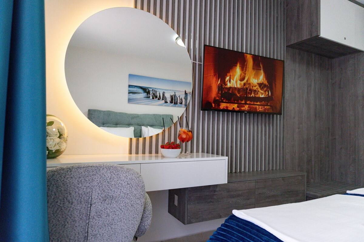Une chambre moderne avec un grand lit, une table de chevet avec un miroir, un téléviseur fixé au mur et une image de cheminée sur le mur.