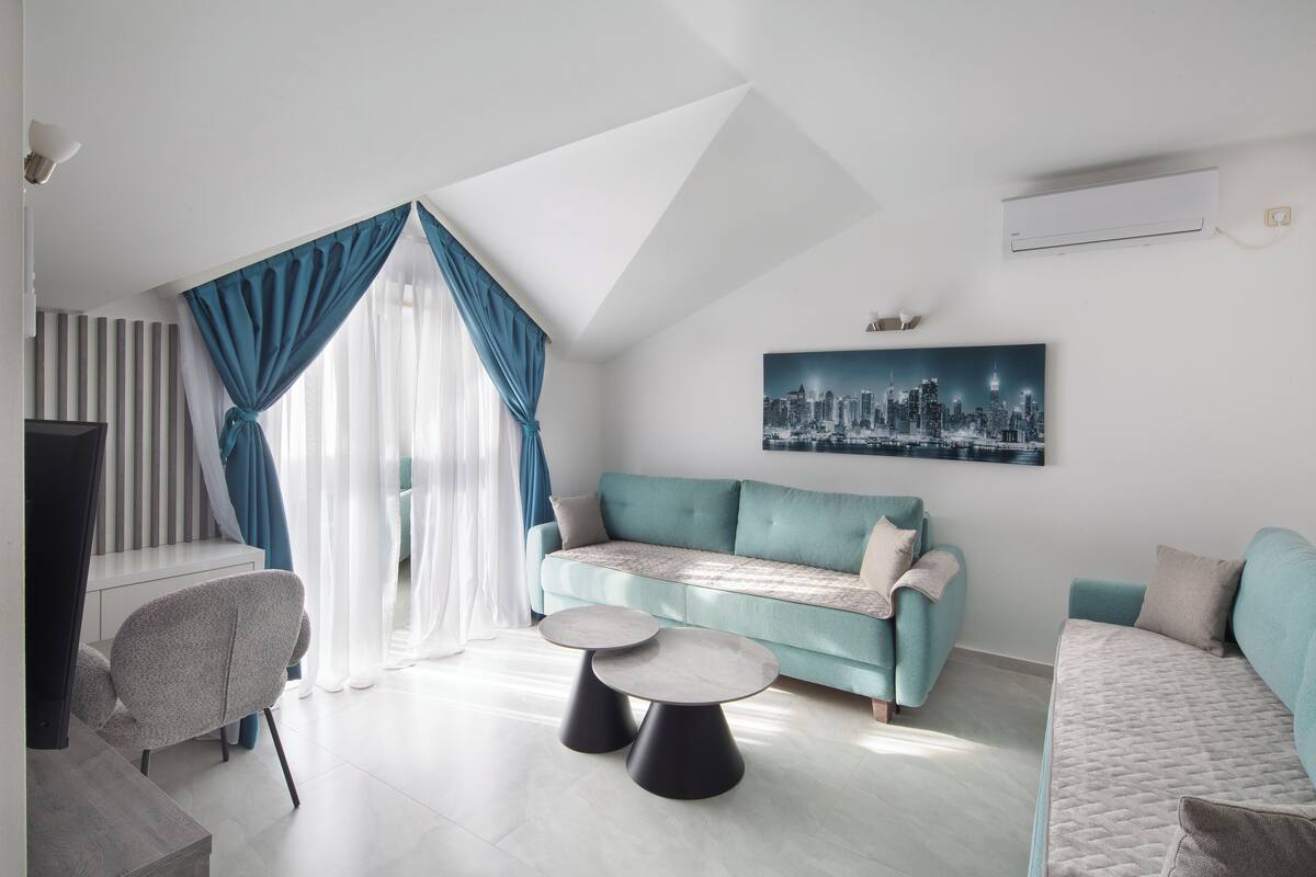 Un salon moderne avec un canapé de couleur turquoise, deux tables rondes, un tableau représentant un paysage urbain et un plafond en pente.