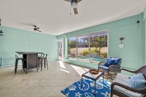 A sunlit patio with a dining table, chairs, and a wicker sofa. A large window offers a view of a swimming pool and greenery. - 3BR/2BA Home | Pool, Ping-Pong & Beach Nearby! (Fort Myers Beach)