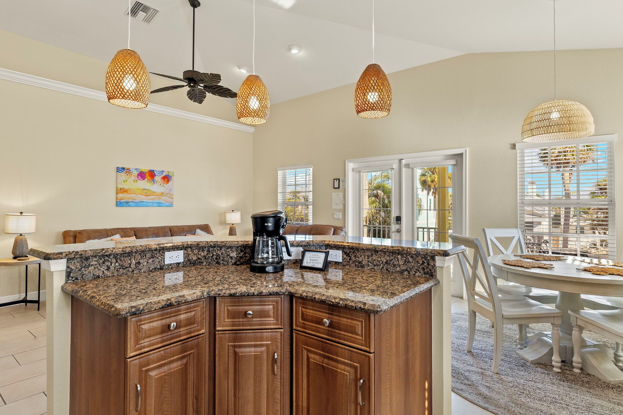 A kitchen with granite countertops, wooden cabinets, and a coffee maker. There are wicker pendant lights, a ceiling fan, and a dining area with a white table and chairs.