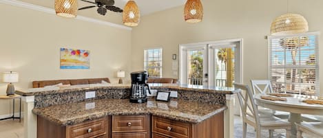 A kitchen with granite countertops, wooden cabinets, and a coffee maker. There are wicker pendant lights, a ceiling fan, and a dining area with a white table and chairs.