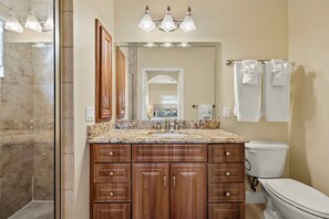 A bathroom with a marble countertop, wooden cabinets, a toilet, and a glass shower enclosure. - 3BR/2BA Home | Pool, Ping-Pong & Beach Nearby! (Fort Myers Beach)