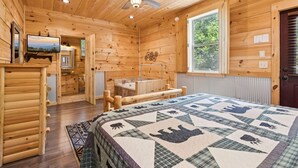 A wooden cabin interior with a bed, a television, a bathroom, and a window with a view of trees.