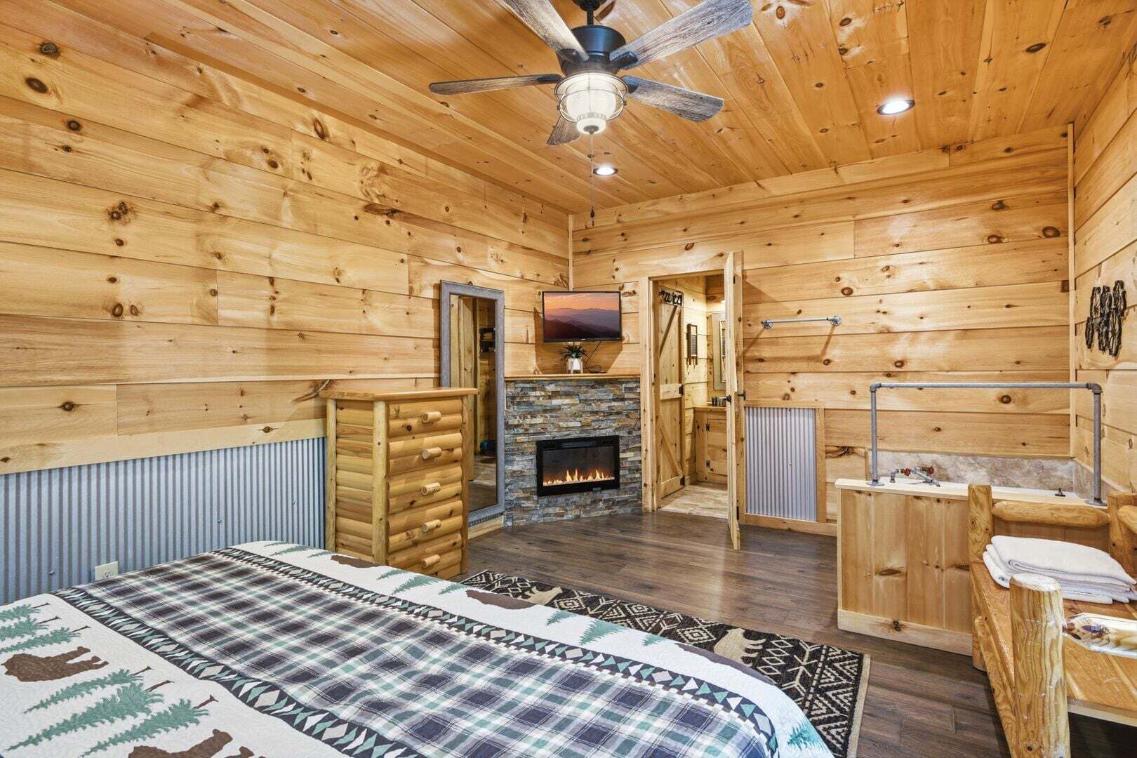 Une chambre dans un chalet en rondins avec une cheminée, un lit recouvert d’une couverture à carreaux, un ventilateur de plafond et un téléviseur fixé au mur.