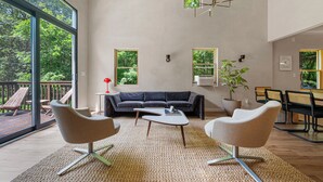 Un salon spacieux doté d’une grande fenêtre, d’un canapé de couleur sombre, de deux fauteuils, d’une table basse et d’un coin repas avec des chaises et une table.