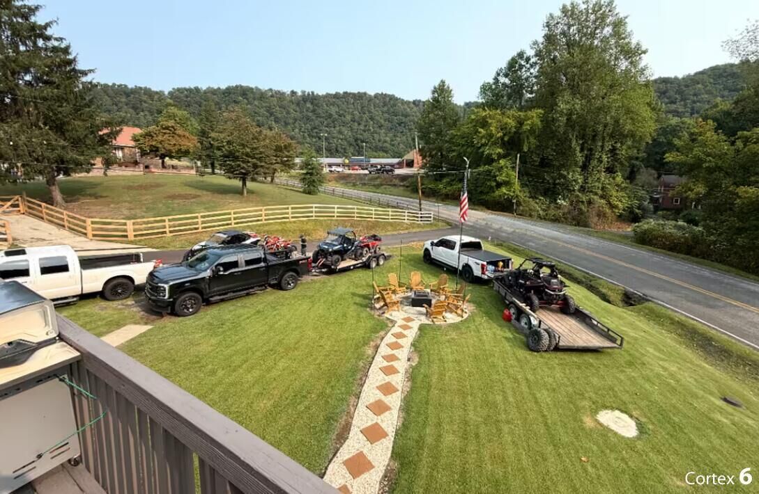 Un césped bien cuidado con un camino pavimentado, una zona de barbacoa y varios vehículos, incluyendo una camioneta, un ATV y un remolque.
