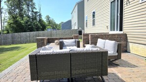 Une terrasse extérieure avec un ensemble de meubles de jardin en osier, une cheminée en briques et une clôture en bois.