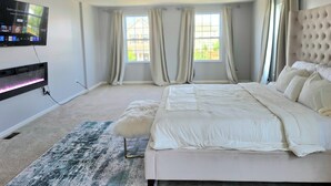 Une chambre à coucher avec un lit, un ventilateur de plafond, un téléviseur fixé au mur et deux fenêtres avec des rideaux.