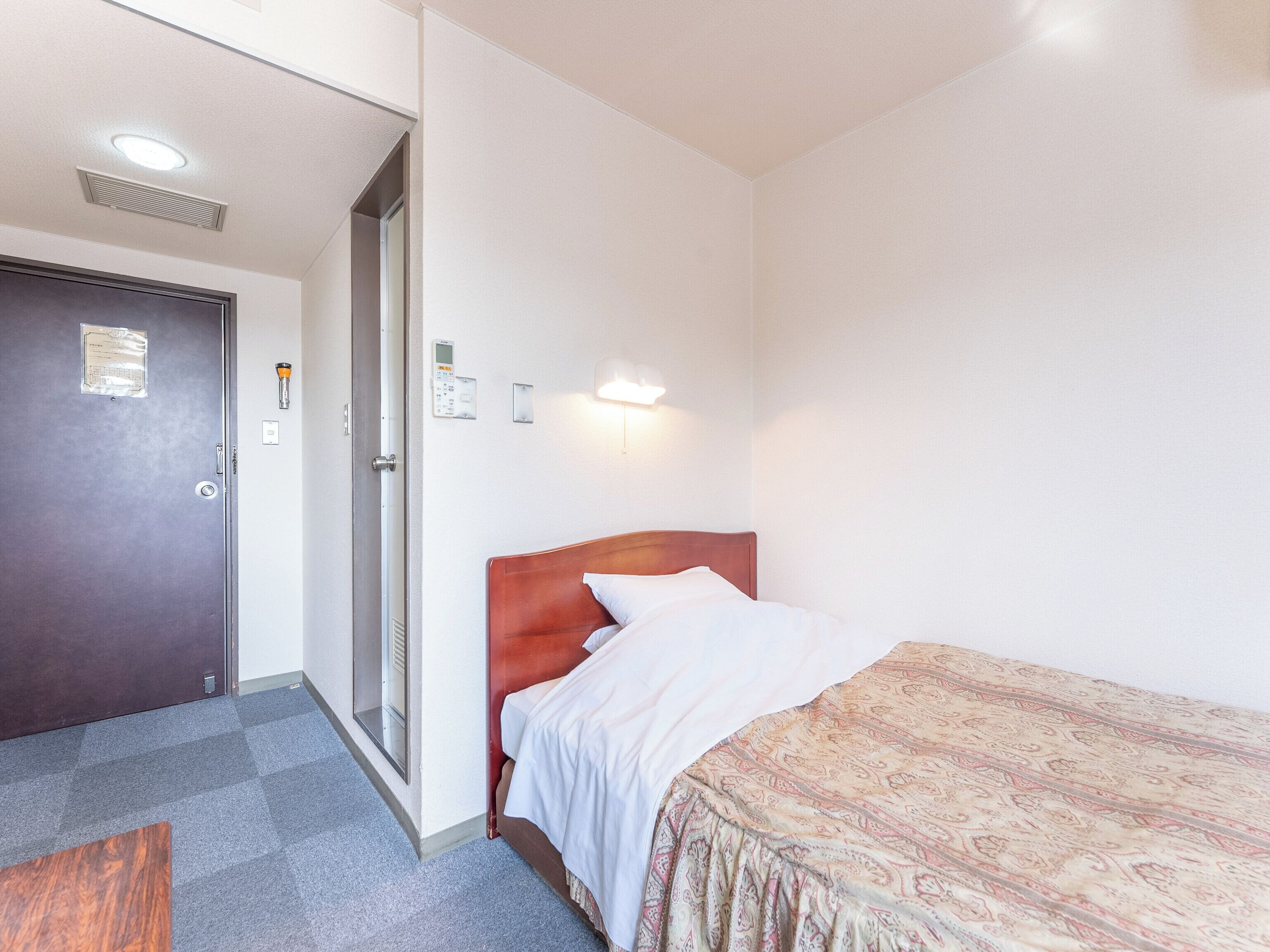 A hotel room with a bed, a door, a light switch, and a thermostat on the wall.