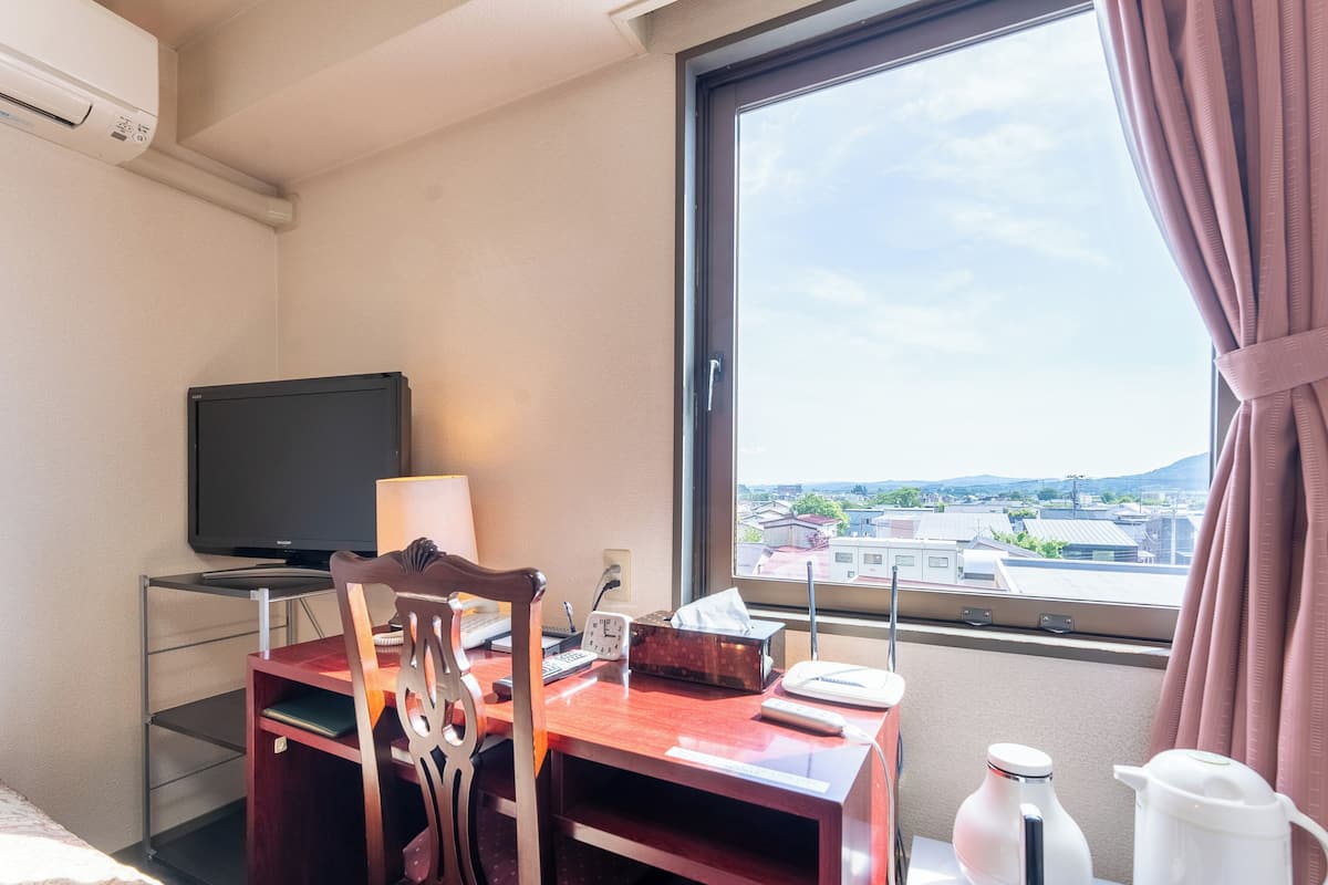 Ein Hotelzimmer mit Schreibtisch, Stuhl, Fernseher, Lampe und einem Fenster mit Blick auf Gebäude und Berge.