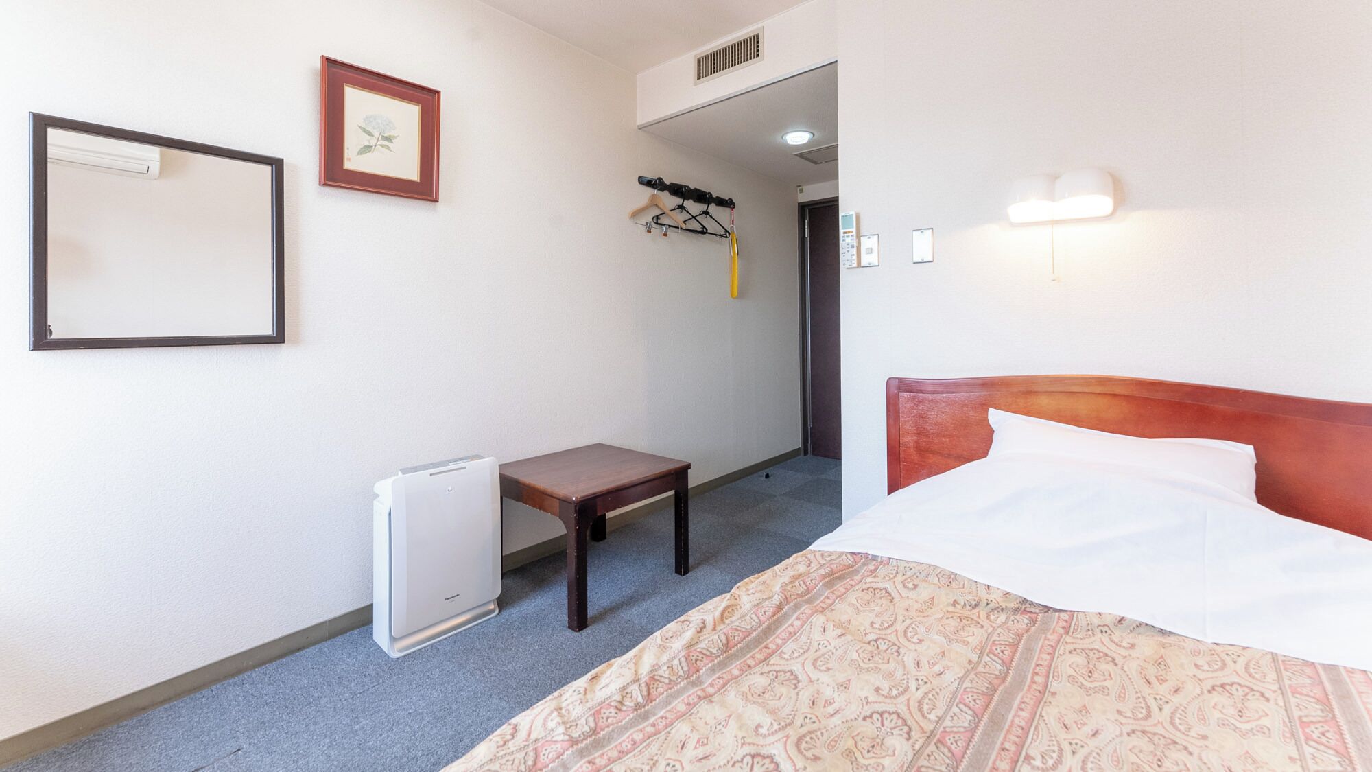 Une chambre d’hôtel avec un lit, une petite table, un radiateur, un porte-manteau et un tableau encadré au mur.