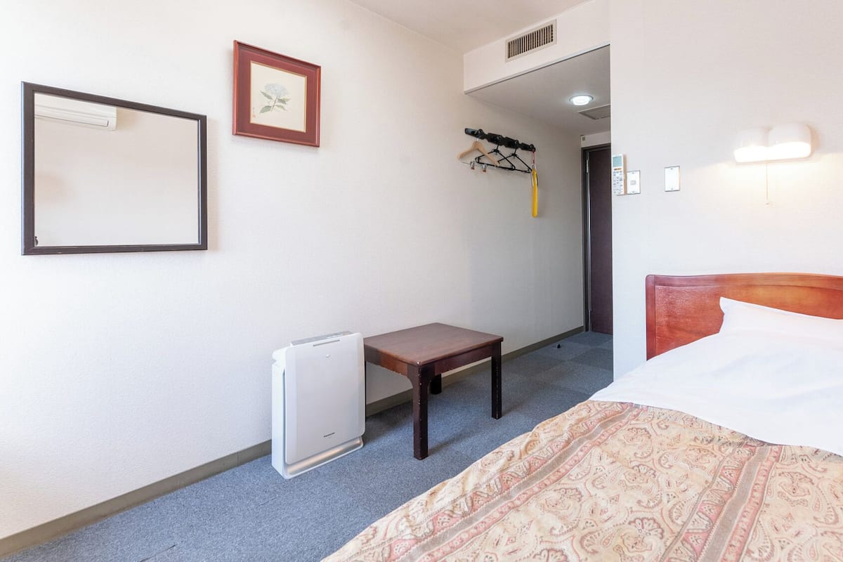Ein Hotelzimmer mit Bett, einem kleinen Tisch, einem Heizkörper, einem Kleiderhaken und einem gerahmten Bild an der Wand.