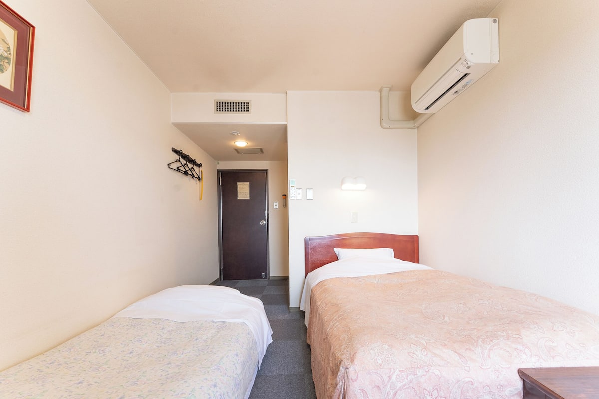 Ein Hotelzimmer mit zwei Einzelbetten, einem Holzkopfende, einem kleinen Tisch, einer Klimaanlage, einer Tür und einer Wandleuchte.