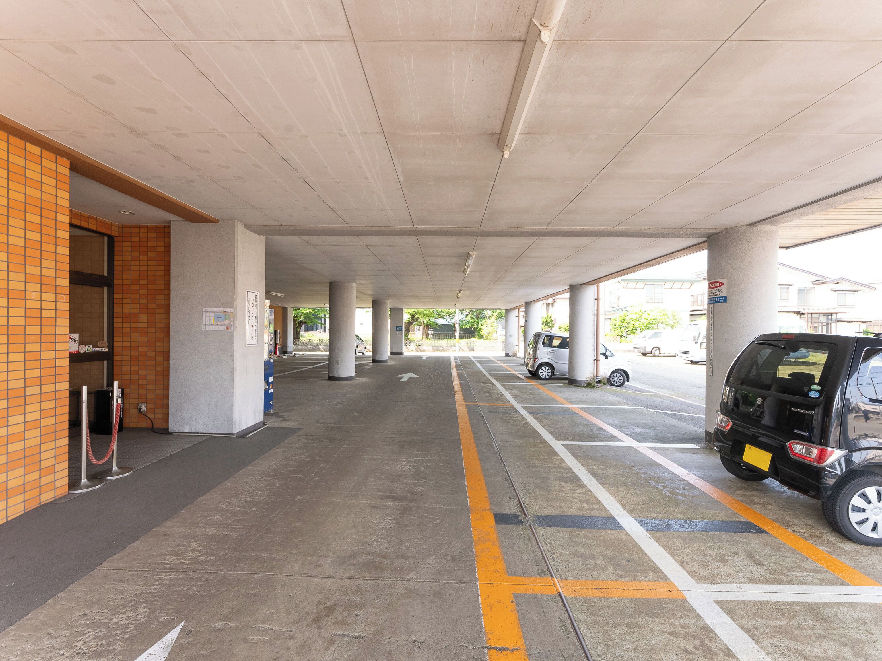 Un área de estacionamiento techada con pilares de hormigón, espacios delimitados y la entrada de un edificio a la izquierda.