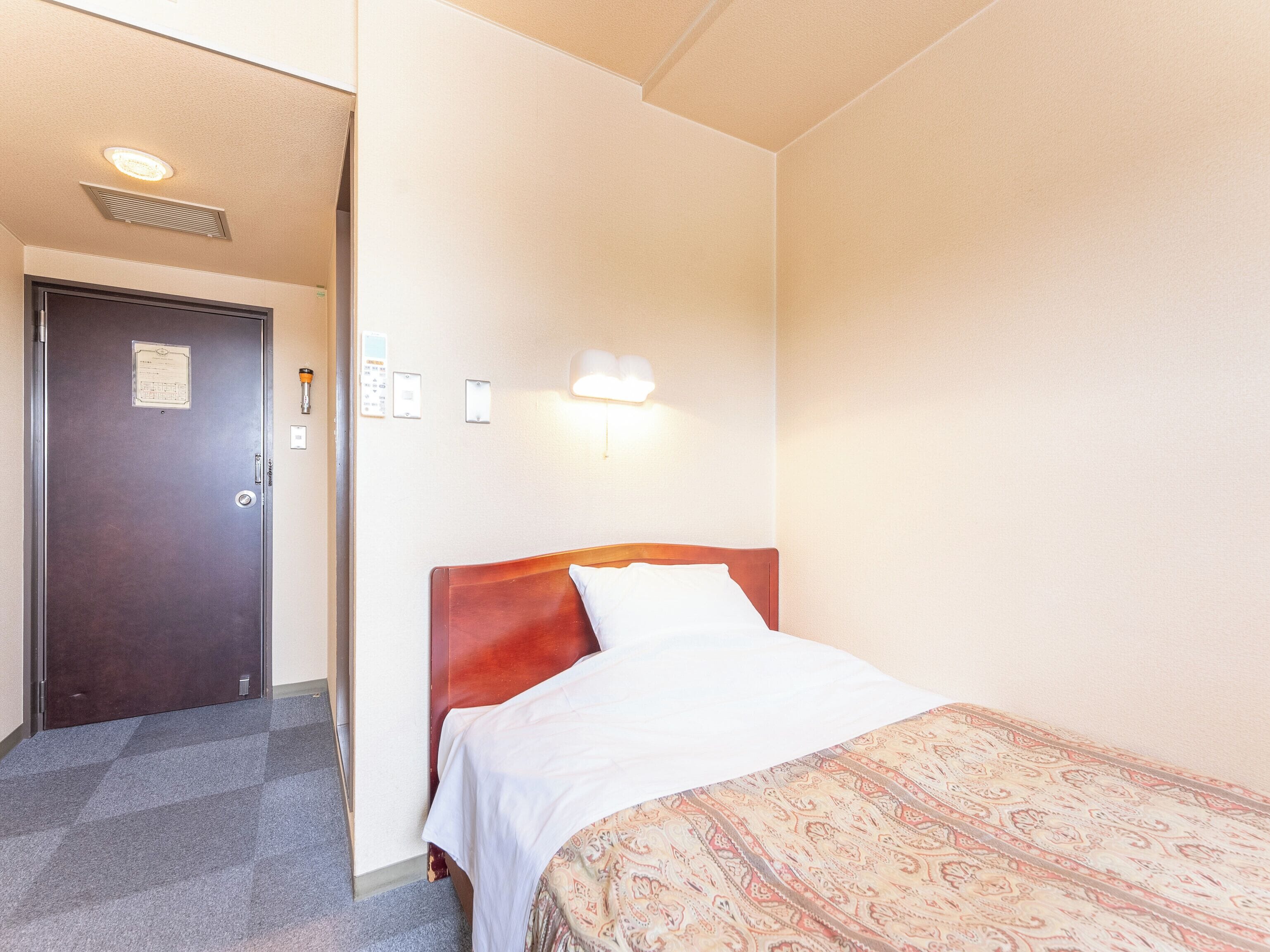 A hotel room with a bed, a door, a light switch, and a wall-mounted light fixture.