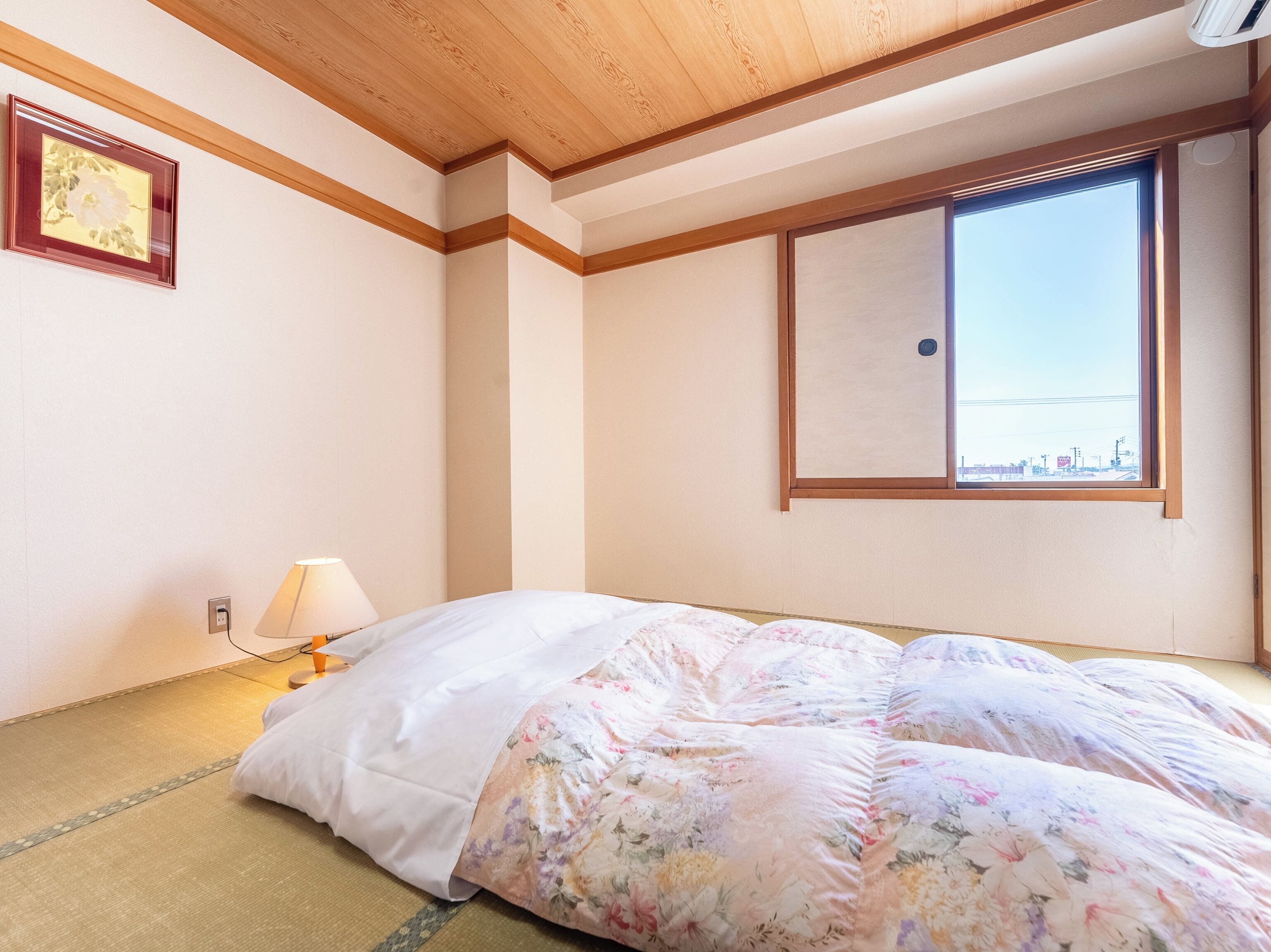 A room with tatami flooring, a bed with a floral comforter, a bedside lamp, a framed picture on the wall, and a window with a view of the outside.