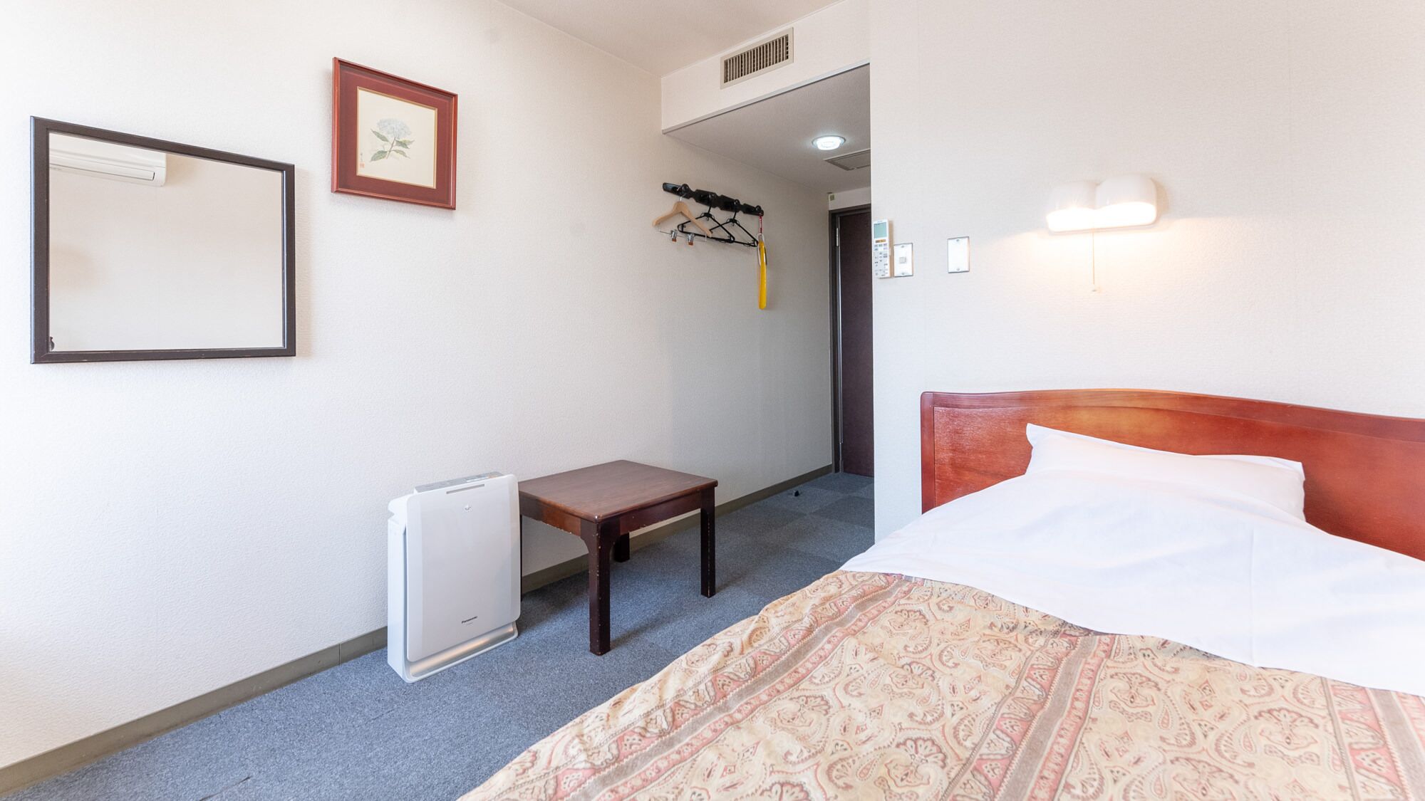 Una habitación de hotel con cama, una mesita, un radiador, un perchero y un cuadro en la pared.