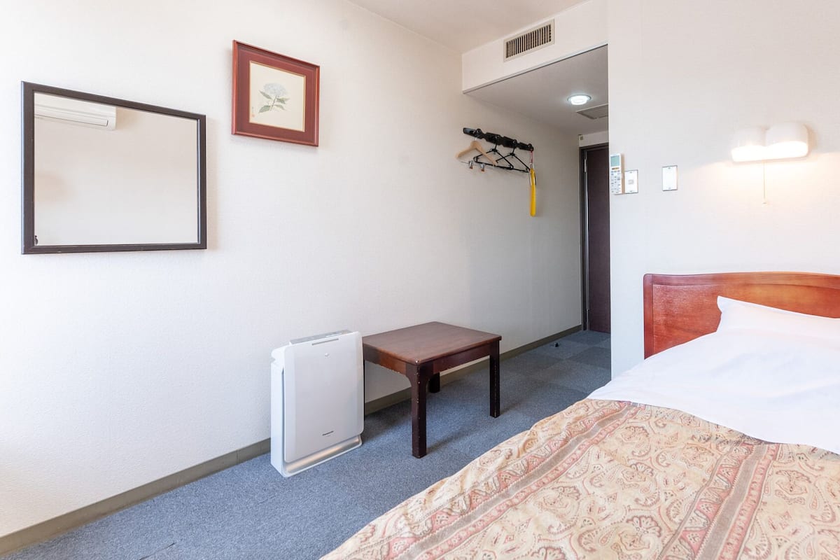 Une chambre d’hôtel avec un lit, une petite table, un radiateur, un porte-manteau et un tableau encadré au mur.
