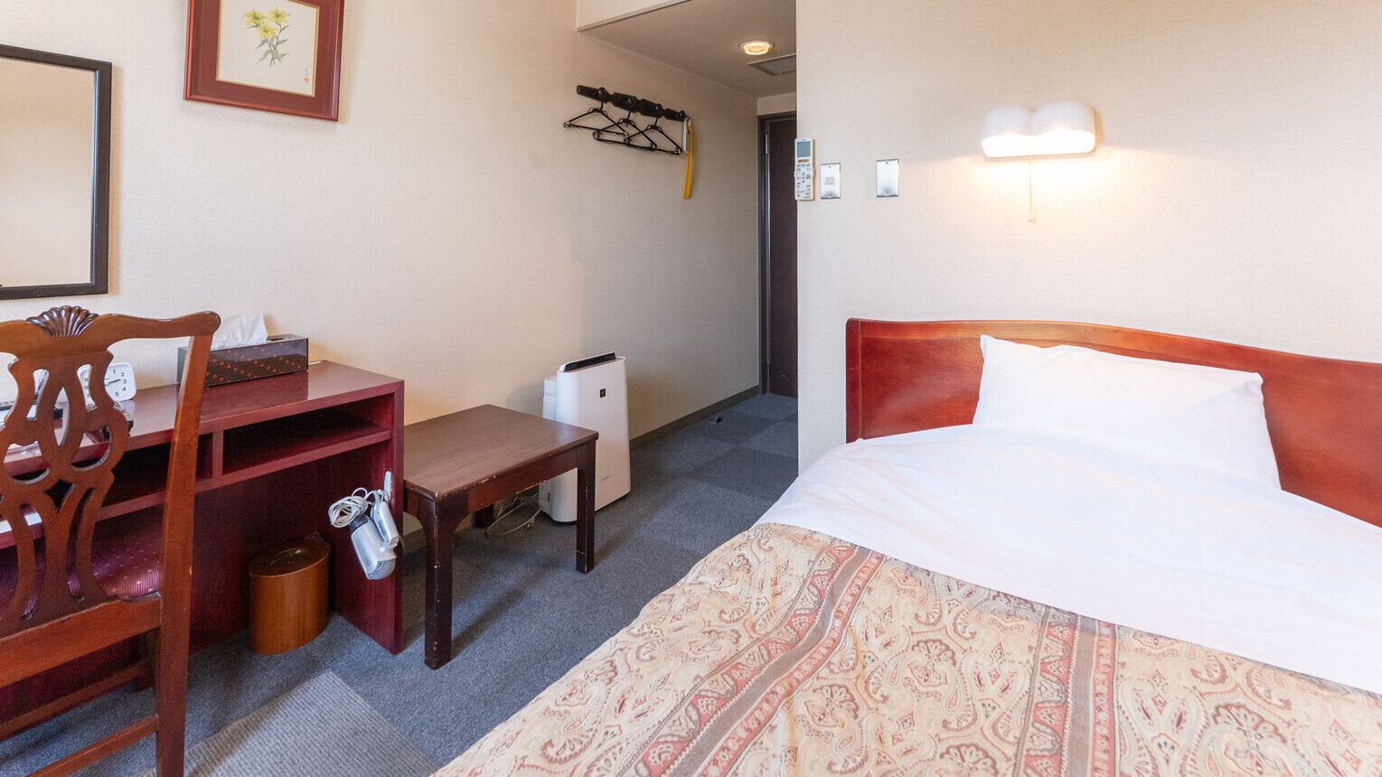 Habitación de hotel con cama, escritorio, silla, cubo de basura y perchero.