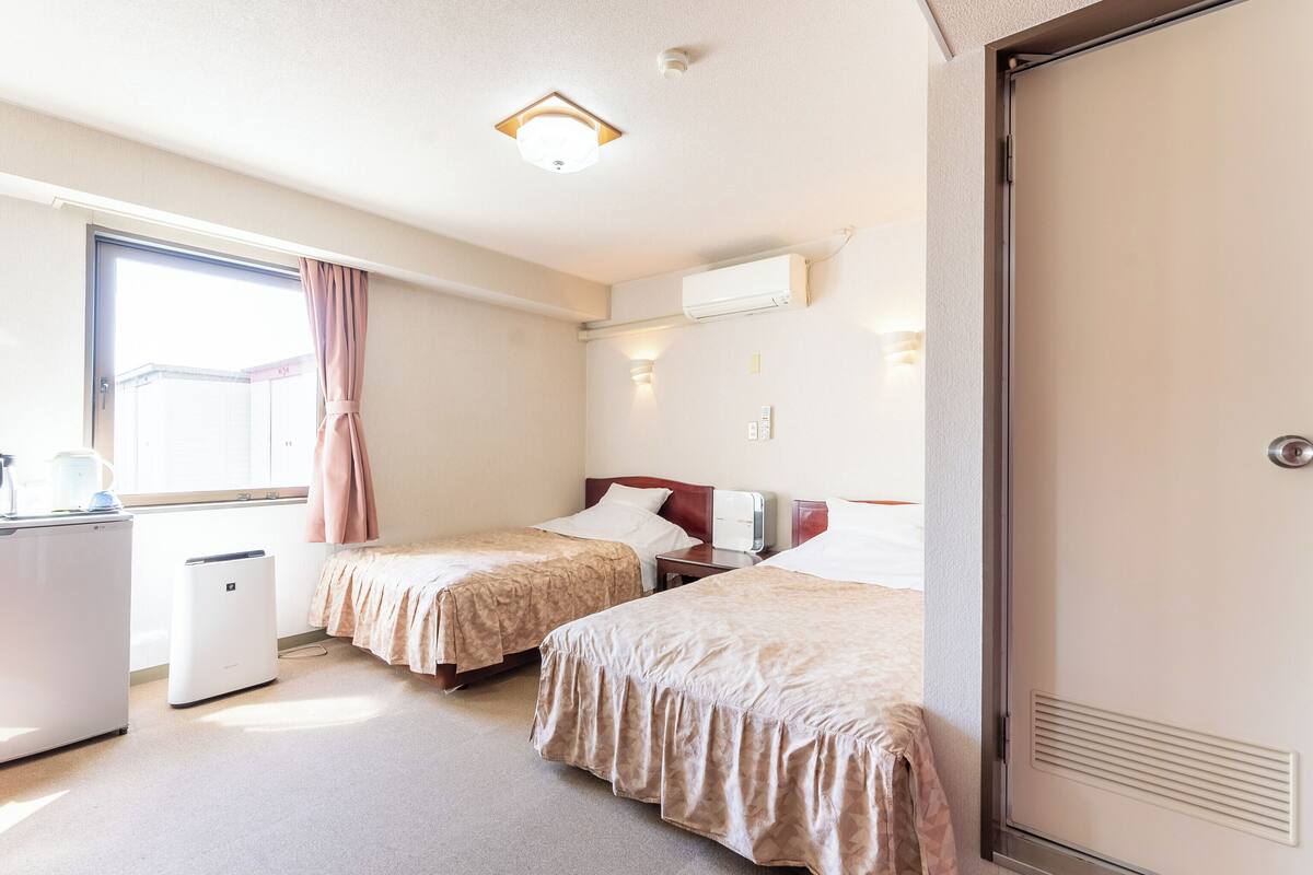 Ein Hotelzimmer mit zwei Betten, einem Schreibtisch, einem Fenster mit Vorhängen, einer Klimaanlage und einer Tür mit Lüftung.