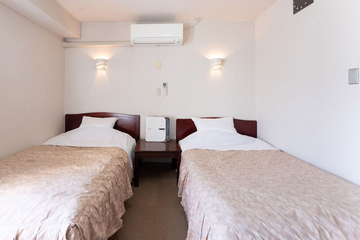 Une chambre d’hôtel avec deux lits, un climatiseur fixé au mur et une table de chevet avec une lampe.