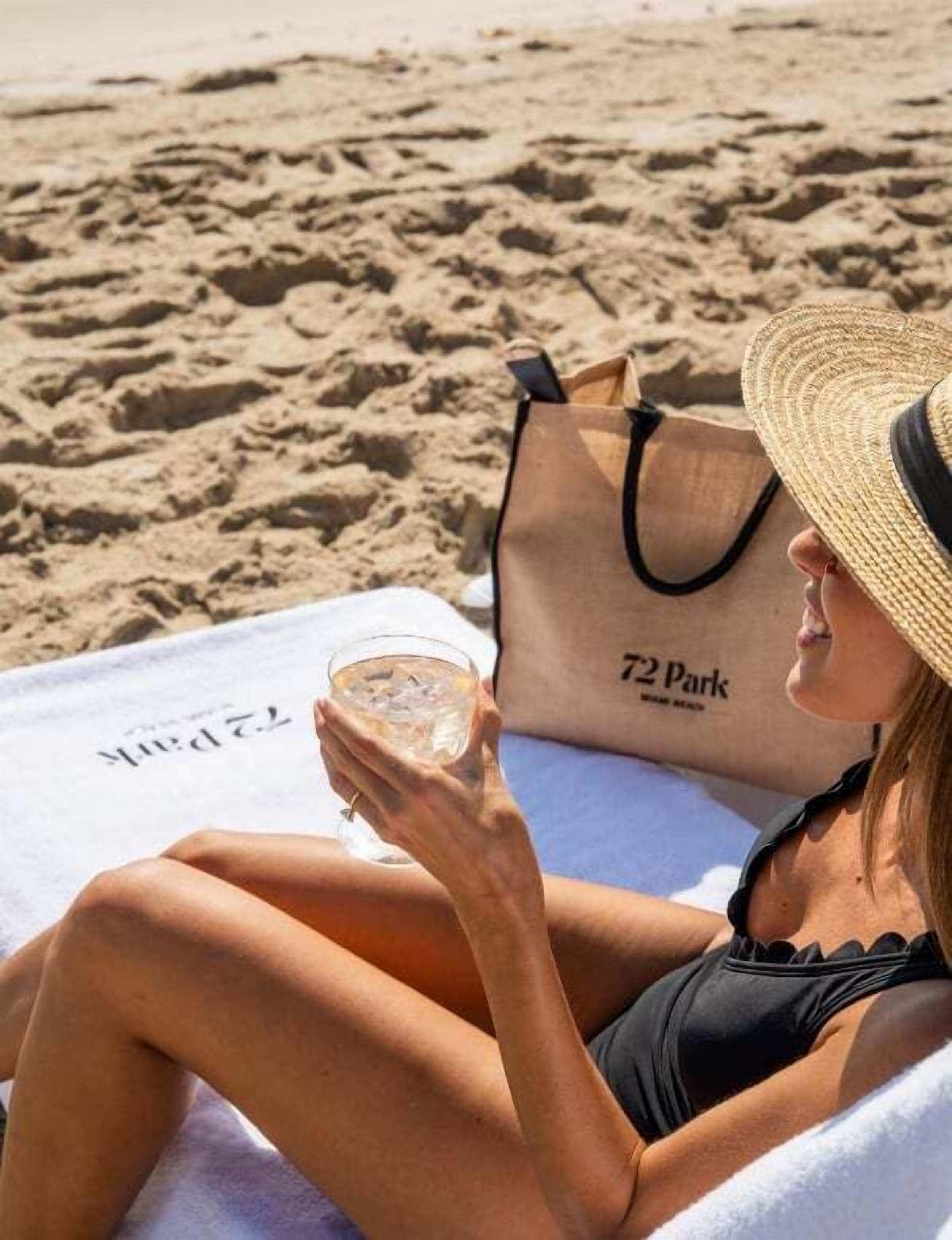 Une personne assise sur une serviette de plage, avec un sac sur lequel on peut lire « 72 Park », et qui tient un verre d’eau.