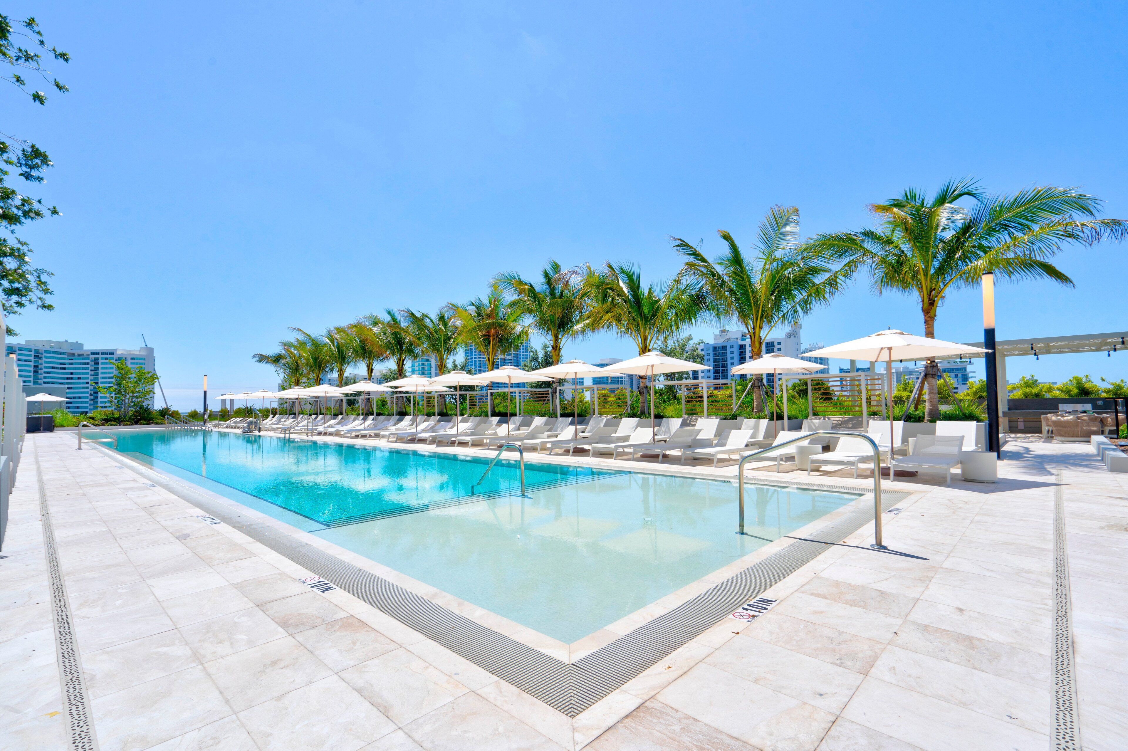 A swimming pool with lounge chairs and umbrellas, surrounded by palm trees and modern buildings.