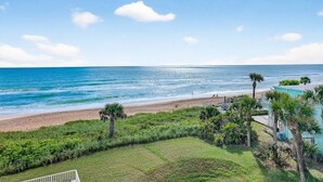A coastal view with a beach, palm trees, and a building with a light blue exterior. - Direct oceanfront corner 2/2 with stunning views, huge balcony, heated pool, and garage parking (Ormond Beach)