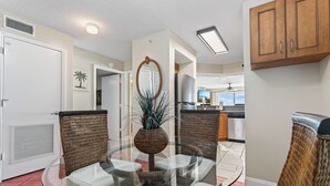 A modern living room with a glass table, wicker chairs, a potted plant, and a kitchen in the background. - Direct oceanfront corner 2/2 with stunning views, huge balcony, heated pool, and garage parking (Ormond Beach)